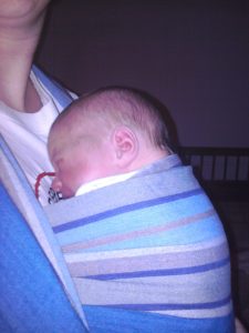 My newborn daughter, aged 3 days, in a stripey bue wrap sling. Her little red face shows how cross she was about being put in the sling.